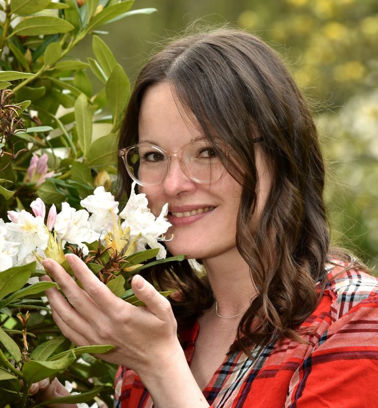 Frau mit Brille hält Blumen in der Hand und lächelt. Hintergrund mit grünen Pflanzen.